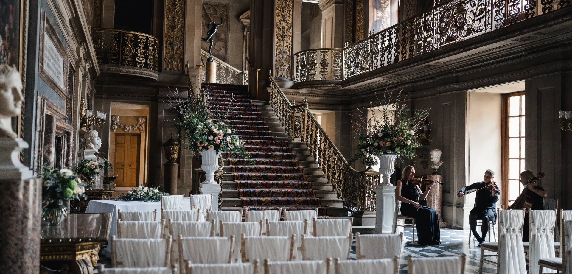 A grand hall with a black and white checkerboard floor, frescos on the walls and ceiling, and a central staircase leading up to a landing.